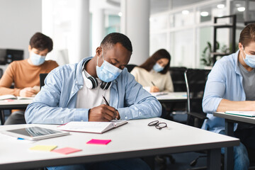 Black male student in mask sitting at desk and writing