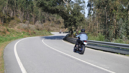 Conjunto de três motards a passear numa estrada de montanha sinuosa - curvas em s duas motas ao longe e uma ao perto - árvore inclinada por cima da estrada