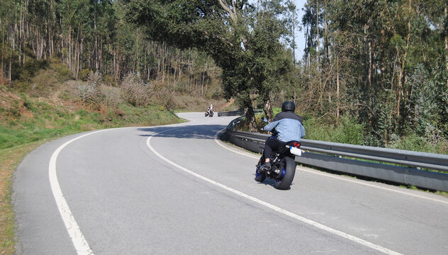 Conjunto de tr&ecirc;s motards a passear numa estrada de montanha sinuosa, curvas em s duas motas ao longe e uma ao perto, &aacute;rvore inclinada por cima da estrada