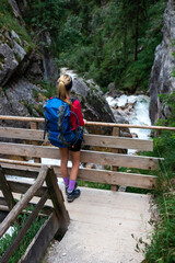 Fototapeta na wymiar Dachstein, Austria: Young attractive blonde woman standing on a bridge in the Silberkarklamm. Beautiful view down the river with wild water during a break from hiking. 