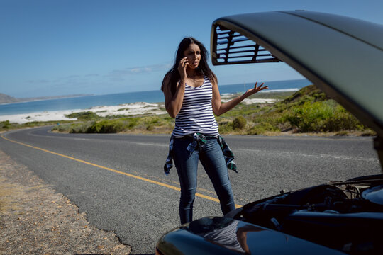 Mixed race woman talking using smartphone standing on road beside broken-down car with open bonnet