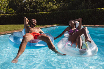 Two diverse male and female friends having fun playing on inflatables in swimming pool
