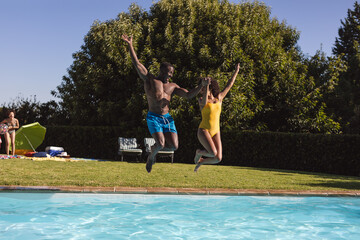 Two diverse male and female friends having fun and jumping into swimming pool