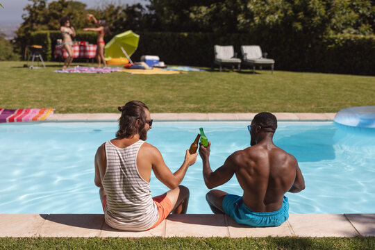 Two diverse male friends drinking beer and sitting at the poolside - Powered by Adobe