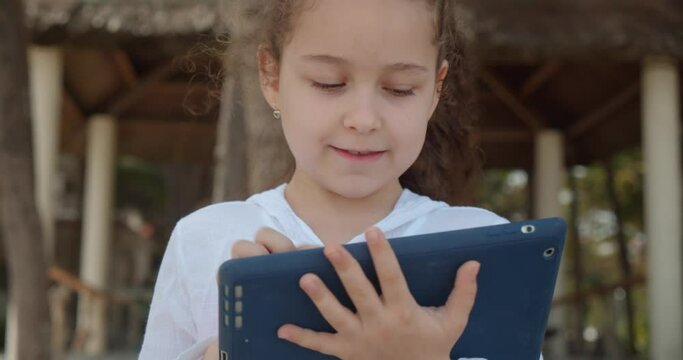 Curious cute girl 7-8 years old is having fun with a digital tablet device watching a cartoon while walking on the beach. The child has a rest plays in the tablet outdoors.