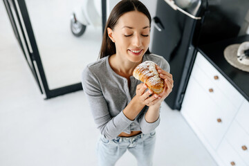 Portrait of rejoicing woman eats tasty croissant at home.