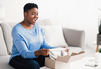 Happy black curvy woman unpacking clothes after online shopping