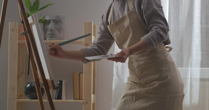 woman painter is drawing picture at home, working in living room, applying dyes on canvas by paintbrush