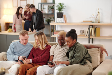 Two young intercultural couples discussing photos in smartphones at home party