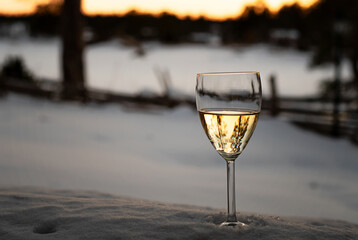 White wine in a wine glass standing in the snow. In the background is view of winter landscape at sunset. Photo taken in Sweden.