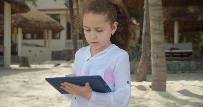 Curious cute girl 7-8 years old is having fun with a digital tablet device watching a cartoon while walking on the beach. The child has a rest plays in the tablet outdoors.
