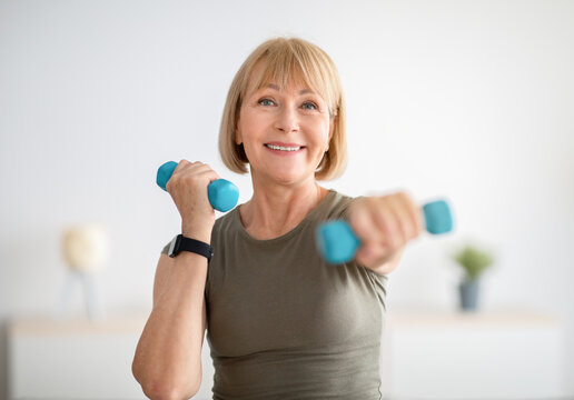 Home training concept. Strong senior woman doing exercises with dumbbells indoors