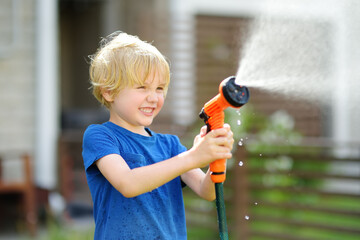 Funny little boy watering lawn and playing with garden hose with sprinkler in sunny backyard. Preschooler child having fun with spray of water.