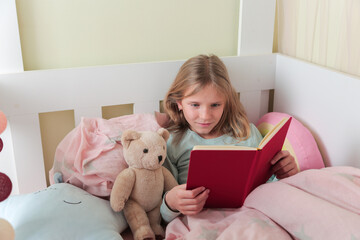 Little blonde girl readingbook . Girl lying on white bed and reading book with her toy bear.