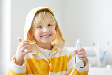 Cute blonde caucasian boy applying cream on his face in bathroom in the morning. Kids care cosmetics. Hygiene for child.