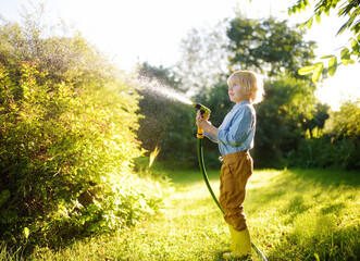 Funny little boy watering plants and playing with garden hose with sprinkler in sunny backyard....