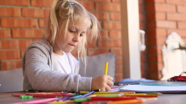 Cute adorable caucasian little blond kid girl enjoy painting with colorful crayons and paper sketchbook at yard table against brick wall. Children creative hobby. Child learning paint at art studio