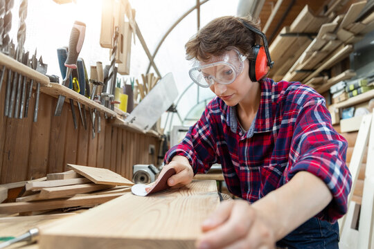 Young attractive 30-40 professional female carpenter looking grinding raw wood with sandpaper in carpentry diy workshop. Feminine women equality concept. Adult girl with male hobby at workbench