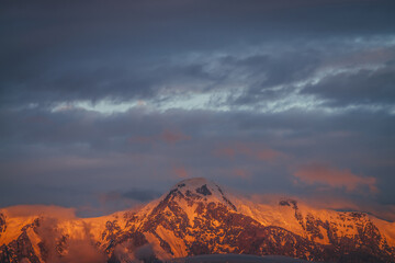 Scenic mountain landscape with great snowy mountains lit by dawn sun among low clouds. Awesome alpine scenery with high mountain pinnacle at sunset or at sunrise. Big glacier on top in orange light.
