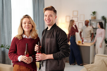 Happy young affectionate couple with bottles of beer looking at you with smiles