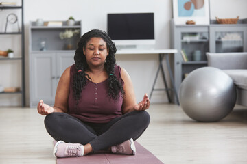Front view full length portrait of curvy African American woman meditating at home while sitting on mat and enjoying home yoga practice, copy space