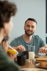 Fototapeta na wymiar Happy young man eating breakfast by served dinner table and looking at his wife