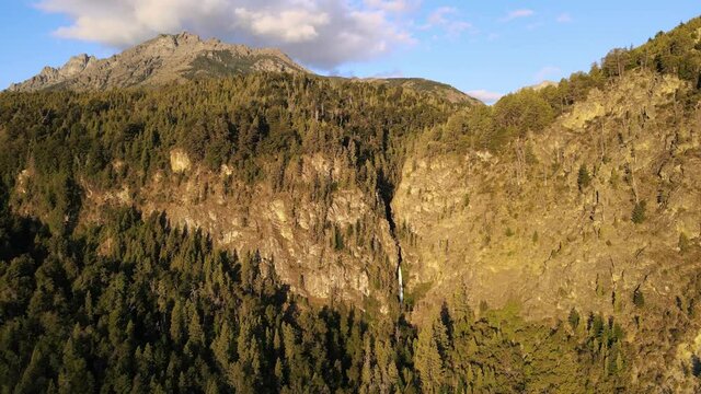 Aerial dolly out of pine tree forest mountains with Corbata Blanca waterfall flowing in background at sunset, Patagonia Argentina
