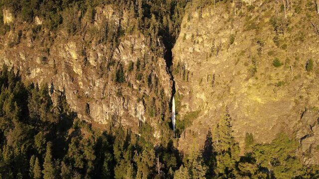 Aerial lowering over Corbata Blanca waterfall hiding between steep mountains with pine trees at sunset, Patagonia Argentina