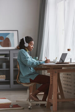 Full length side view at young African American woman studying at home or working at desk while wearing headphones