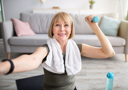 Joyful mature woman taking selfie while exercising with dumbbells at home