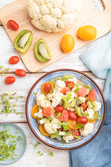 Vegetarian salad of cauliflower cabbage, kiwi, tomatoes, microgreen sprouts on gray wooden background. Top view, close up.