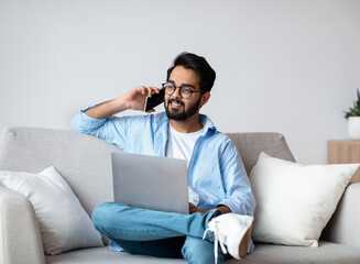 Obraz premium Remote Job. Eastern Man Talking On Cellphone And Using Laptop At Home