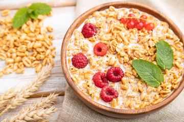 Wheat flakes porridge with milk, raspberry and currant in wooden bowl on white wooden background. Side view, selective focus.
