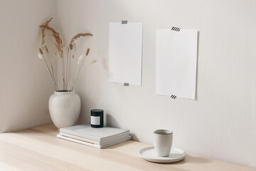 Breakfast still life. Cup of coffee, candle and books on wooden desk, table. Empty paper sheets and posters mockups taped on white wall.Vase with dry grass.Scandinavian working space, home office.