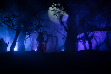 Horror Halloween decorated conceptual image. Alone girl with the light in the forest at night. Silhouette of girl standing between trees with surreal light on background. Selective focus.
