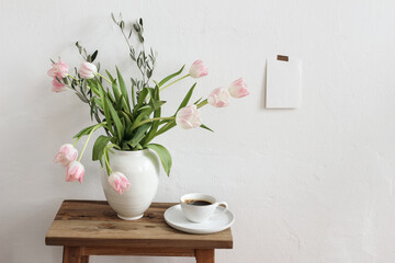 Easter, spring still life scene. Cup of coffee and floral bouquet in ceramic vase on wooden bench. Pink tulips flowers, olive tree branches on table.Blank greeting card mockup taped on white wall.