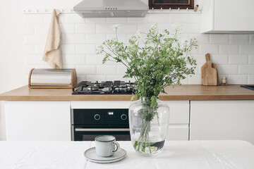 Cup of coffee, vase with cow parsley on white linen table cloth. Modern eclectic kitchen interior....