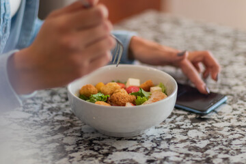 Close up of a young brunette woman eating a salad in her kitchen. She is using her phone