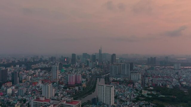Fly in towards Ho chi Minh City center before dawn with colorful sky. Key skyscrapers of the city are in the middle of frame with reflections in the canal flanked by modern residential towers