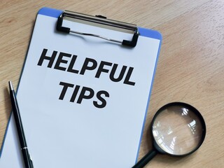 Phrase HELPFUL TIPS written on paper clipboard with pen and magnifying glass. Business concept.