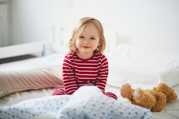 Happy toddler girl in striped red and white pajamas sitting on bed right after awaking - obrazy, fototapety, plakaty