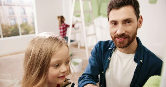 Caring father paints wall in room with his little daughter, helping by holding paint roller. Concept of father and child on housework.
