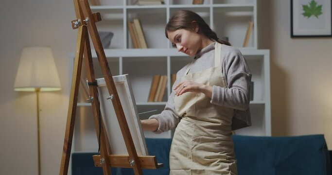 young cheerful woman is drawing on canvas on easel in apartment at evening, hobby and favorite pastime
