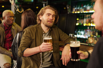 Young bearded man in casualwear having beer with his friend by bar counter