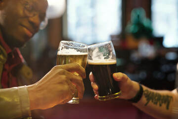Hands of two young intercultural men clinking with glasses of beer in the pub