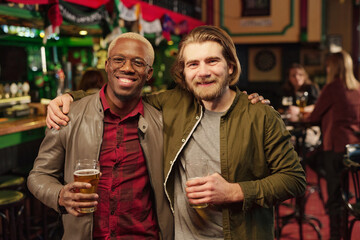 Two young cheerful intercultural men looking at you while toasting with beer