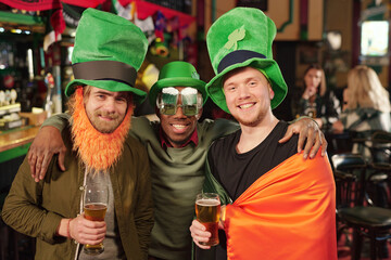 Three happy young intercultural men in green hats toasting with glasses of beer