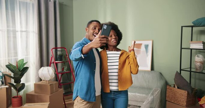Young happy african american couple in new apartment talking to friends over video call using smartphone. New homeowners are blogging.