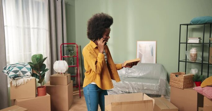 Young beautiful african american woman moved into new apartment and unpacks boxes with things while talking on cell phone with friend.