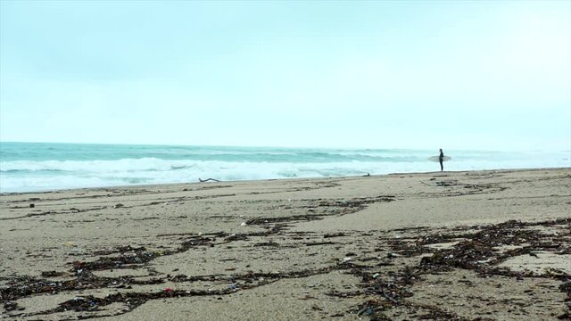 Solo surfer standing on beach with waves crashing in super slow motion. Extreme wide shot with subject tiny in frame. 

Filmed in France on Biaritz beach.